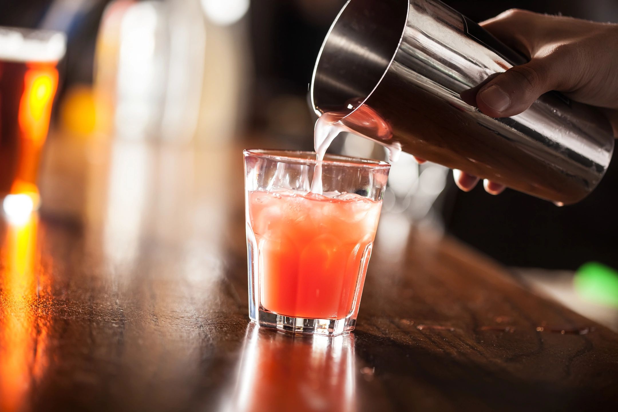 A vibrant pink cocktail being poured into a glass on a wooden bar.
